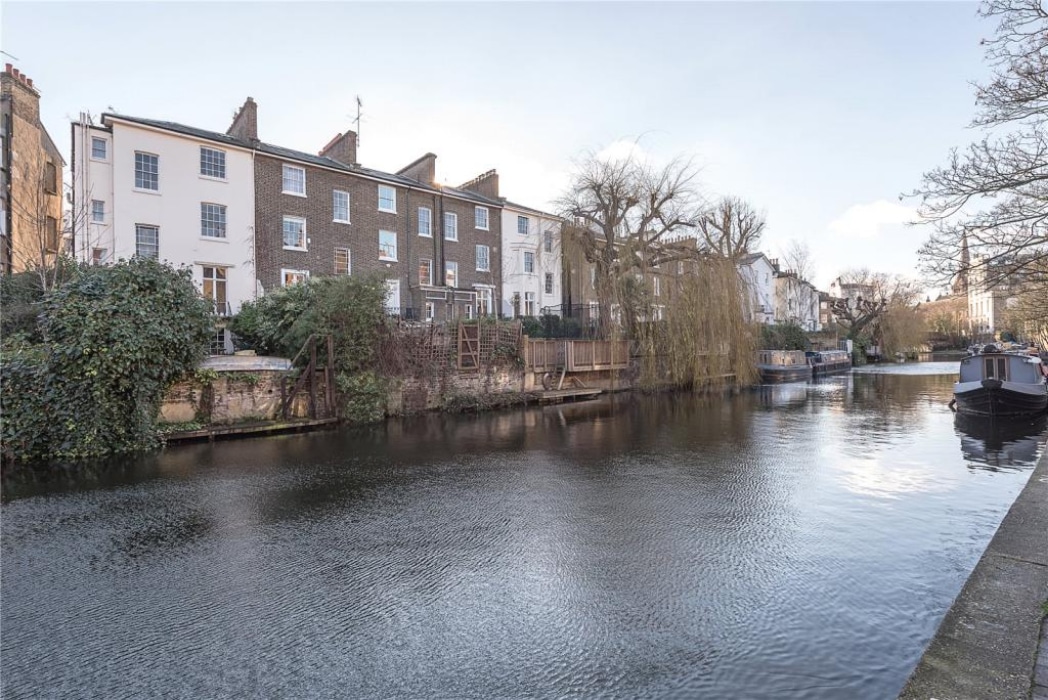 Row of terraced houses with gardens along a canal, with moored boats and overhanging trees reflected in the water.