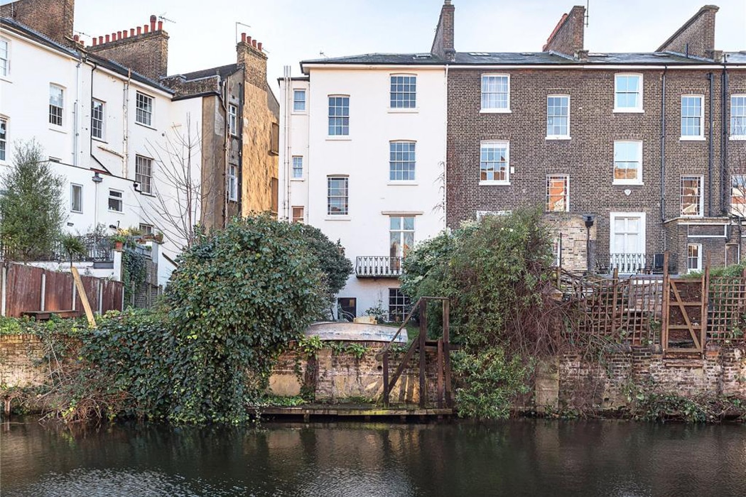 Row of residential buildings with brick and white facades behind overgrown vegetation and a small canal with calm water in the foreground.