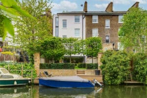 Three-story brick and stucco townhouse with riverside garden, terrace, and private dock holding two boats, surrounded by trees and greenery.