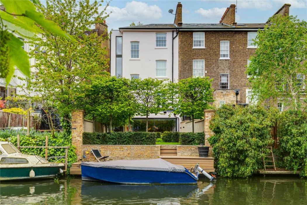 Three-story brick and stucco townhouse with riverside garden, terrace, and private dock holding two boats, surrounded by trees and greenery.