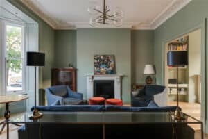 A living room with sage green walls, a fireplace, two blue armchairs, a dark sofa, two black floor lamps, and a painting above the mantel.