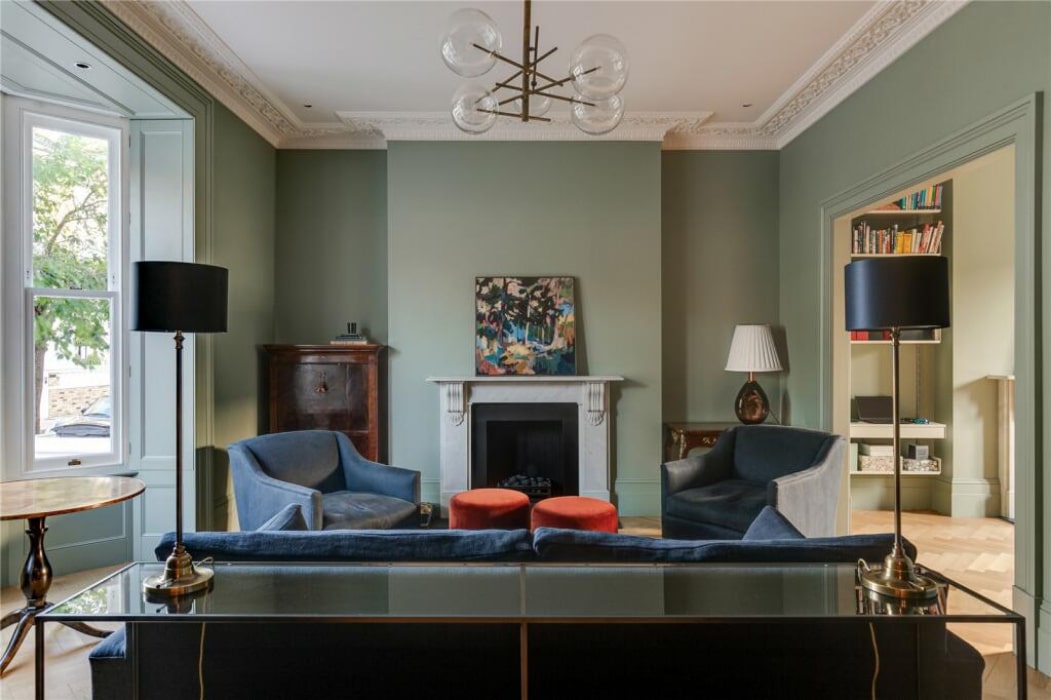 A living room with sage green walls, a fireplace, two blue armchairs, a dark sofa, two black floor lamps, and a painting above the mantel.