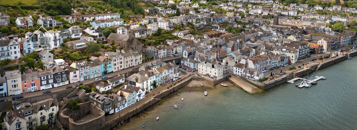 Aerial view of a coastal town with rows of pastel-colored houses, narrow streets, small boats on the water, and a harbor with docks.