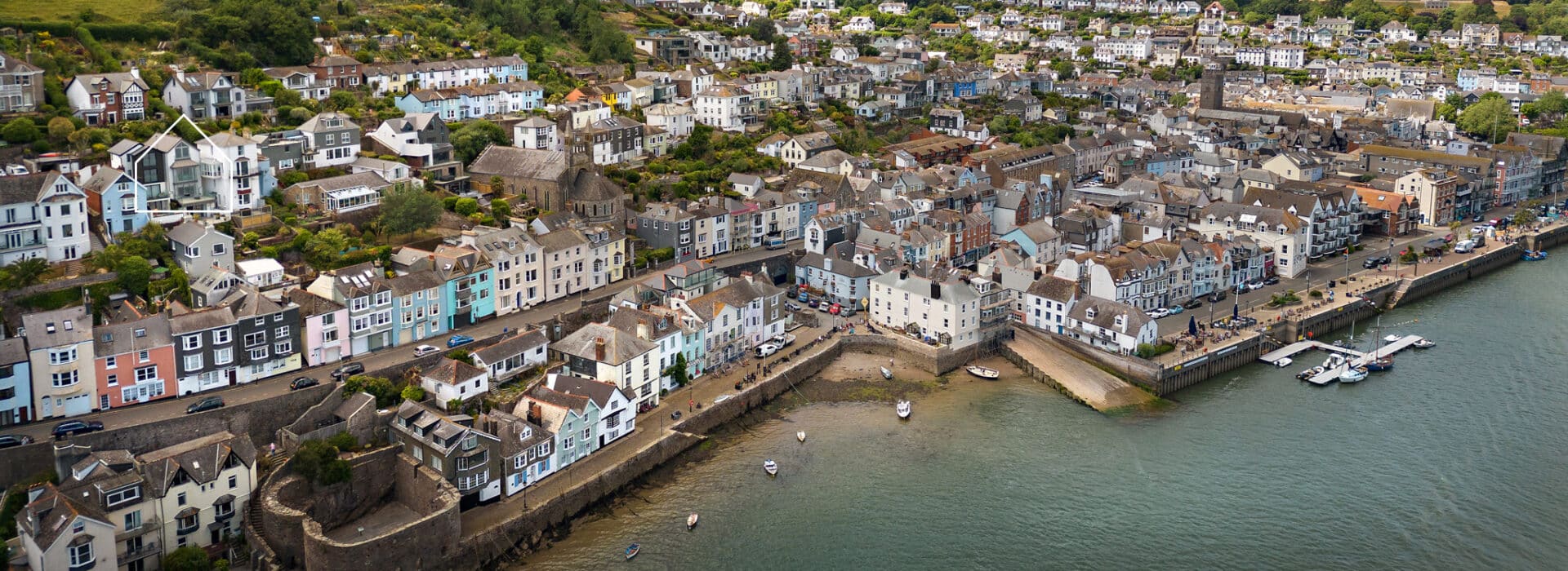 Aerial view of a coastal town with rows of pastel-colored houses, narrow streets, small boats on the water, and a harbor with docks.