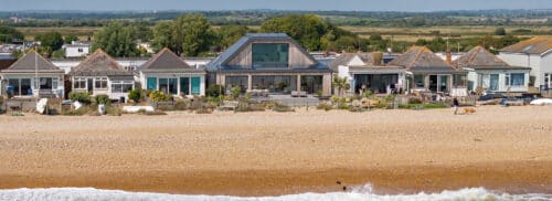 Row of modern beach houses with large windows and terraces facing a pebble beach, with grassy landscape in the background.