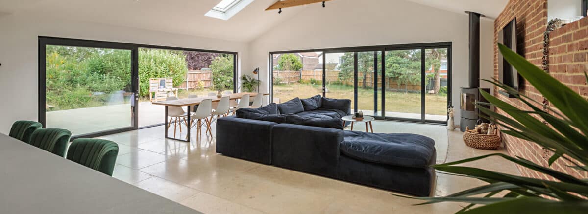Modern open-plan living and dining area with large windows, navy sectional sofa, dining table with white chairs, and views of a green backyard.