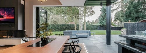 Modern indoor dining area opens to a patio with outdoor seating, overlooking a spacious green lawn and trees.