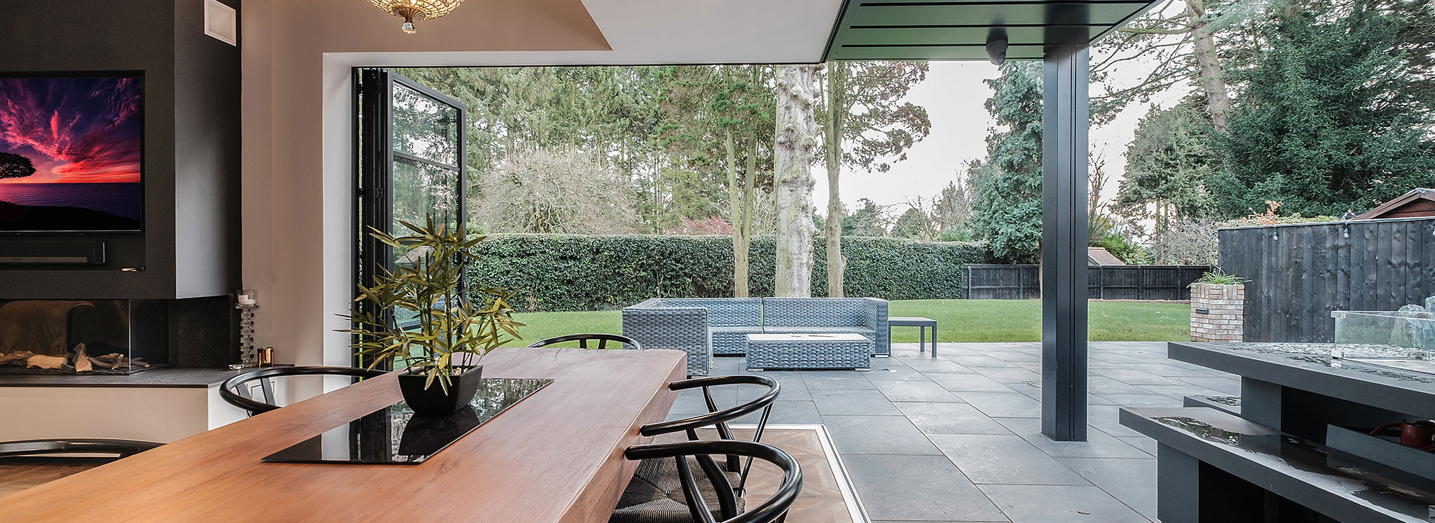 Modern indoor dining area opens to a patio with outdoor seating, overlooking a spacious green lawn and trees.