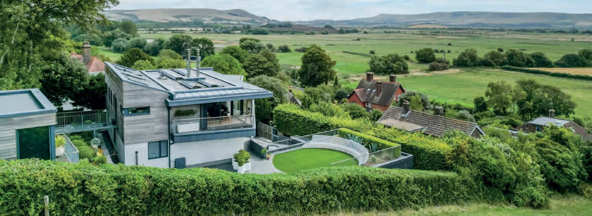 Modern house with large windows and rooftop solar panels overlooks green fields and distant hills, surrounded by trees and hedges.