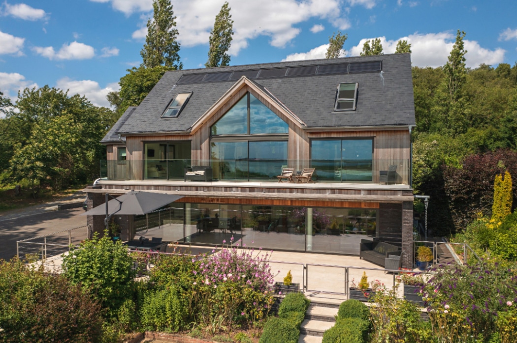 A modern two-storey house with large glass windows, a sloping roof, solar panels, and a spacious terrace, surrounded by greenery and a landscaped garden.