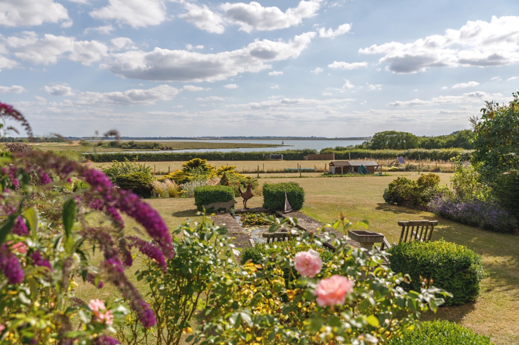 A landscaped garden with pink and purple flowers, wooden chairs, and outdoor sculptures, overlooking open fields and a distant body of water under a partly cloudy sky.