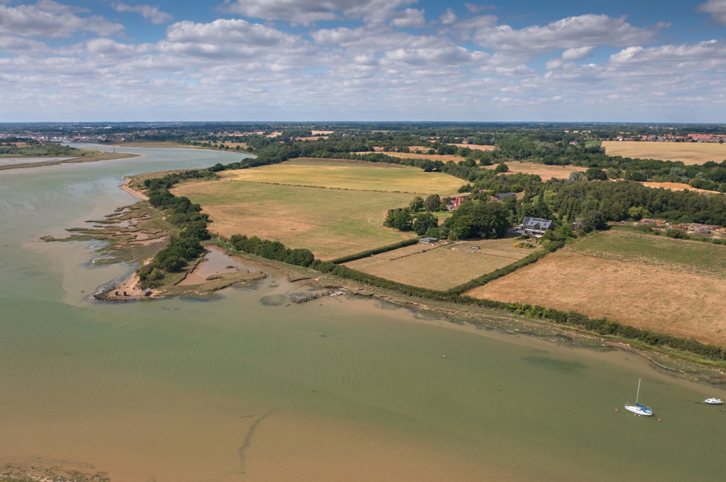 Aerial view of a river estuary with grassy fields, trees, and a few buildings along the shore under a partly cloudy sky.