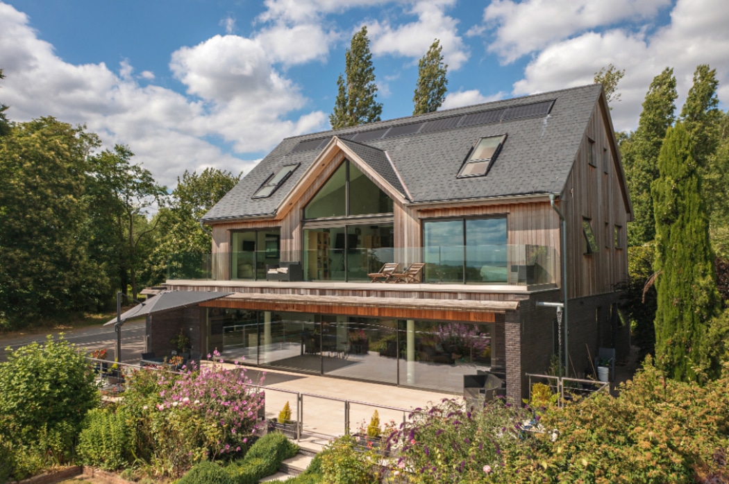 A modern two-storey house with large glass windows, a sloped roof with solar panels, a wide balcony, and landscaped greenery at the front.