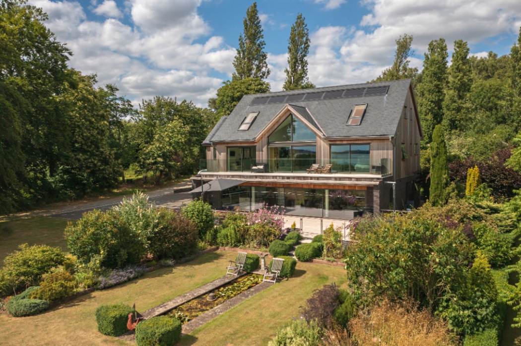 Modern two-storey house with large glass windows, surrounded by landscaped gardens, a pond, and outdoor seating, set amid trees under a partly cloudy sky.