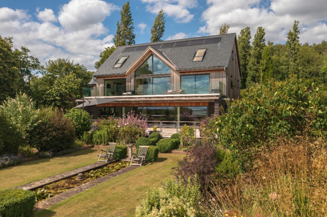 Modern two-storey house with large glass windows, solar panels on the roof, and a landscaped garden with a pond and outdoor seating in the foreground.