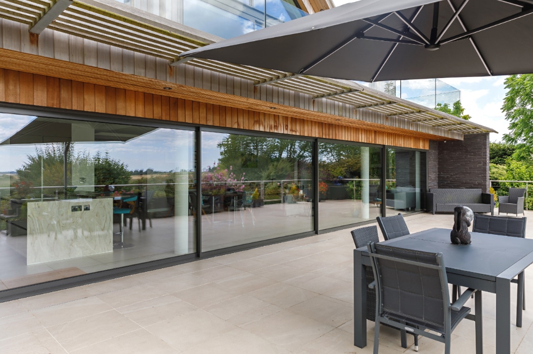 Modern outdoor patio with large sliding glass doors, dining table and chairs, parasol, and a view of greenery reflected in the windows.
