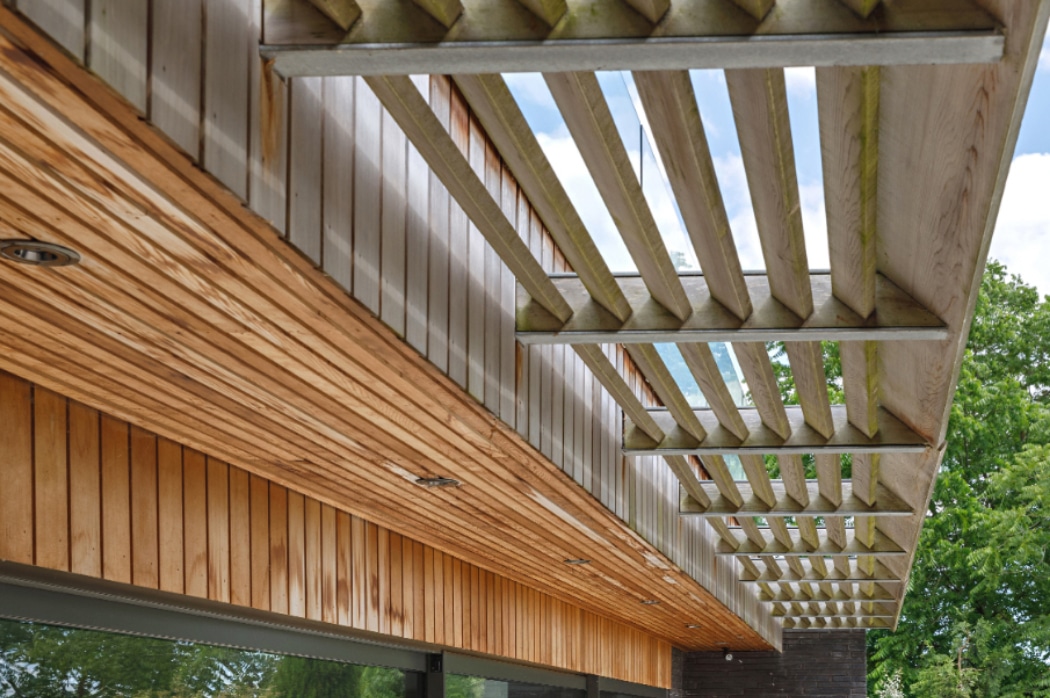 Wooden slatted pergola structure extends from the roof of a modern building, with trees and sky visible in the background.