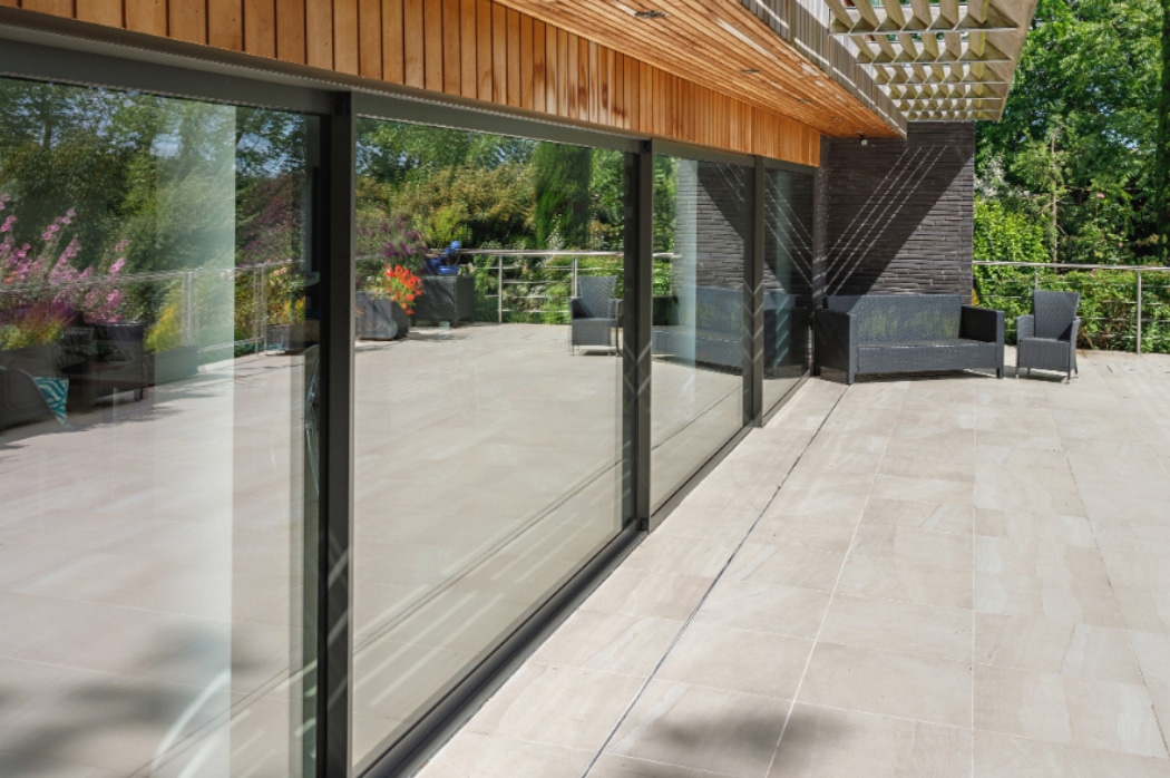 Spacious outdoor patio with light-coloured tile flooring, glass sliding doors, wicker chairs, and glass railing overlooking greenery.