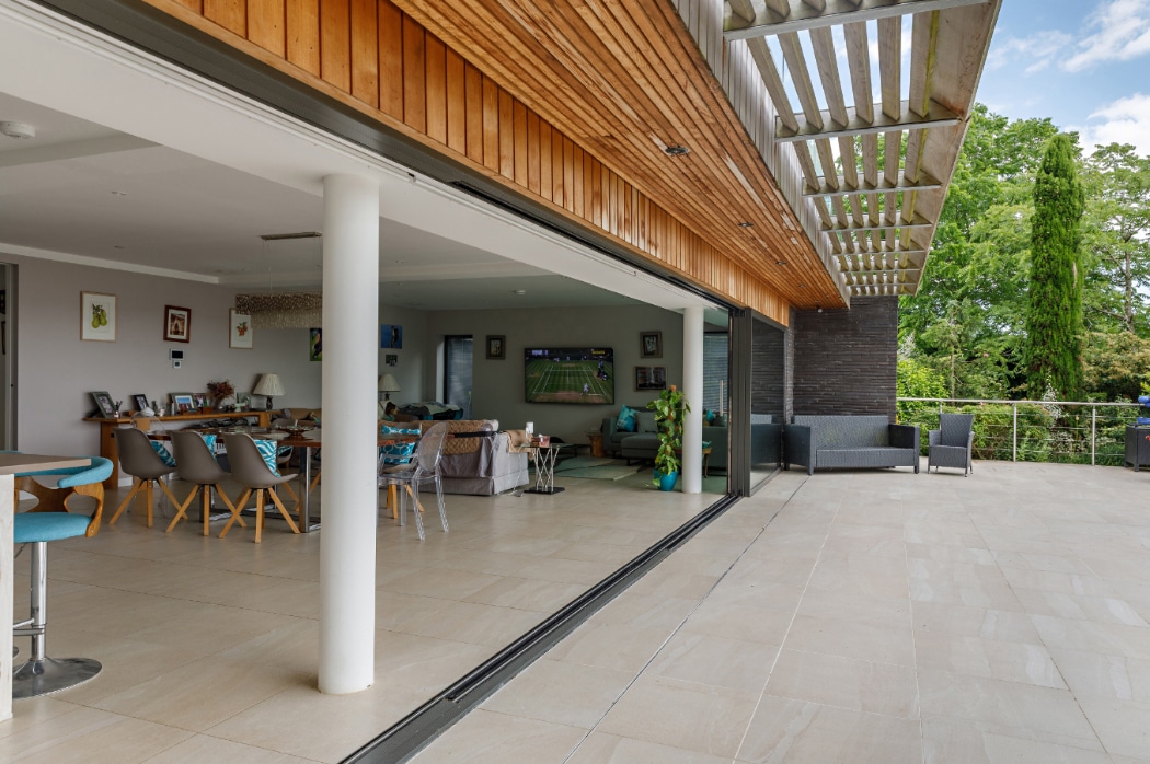 Spacious modern open-plan living area with large sliding glass doors leading to a tiled outdoor patio and seating area, surrounded by greenery.