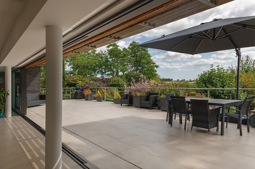 Spacious outdoor terrace with tiled flooring, black dining set, parasol, potted plants, and views of trees and fields under a partly cloudy sky.