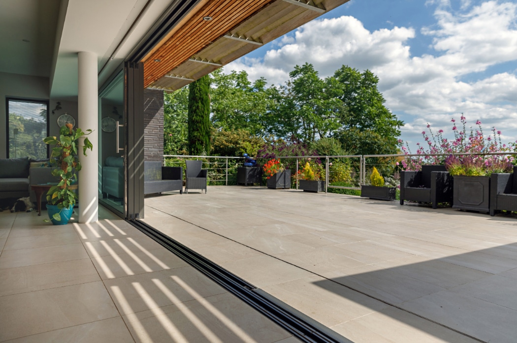 Spacious modern patio with open sliding glass doors, tiled flooring, potted plants, outdoor seating, and a view of trees under a partly cloudy sky.