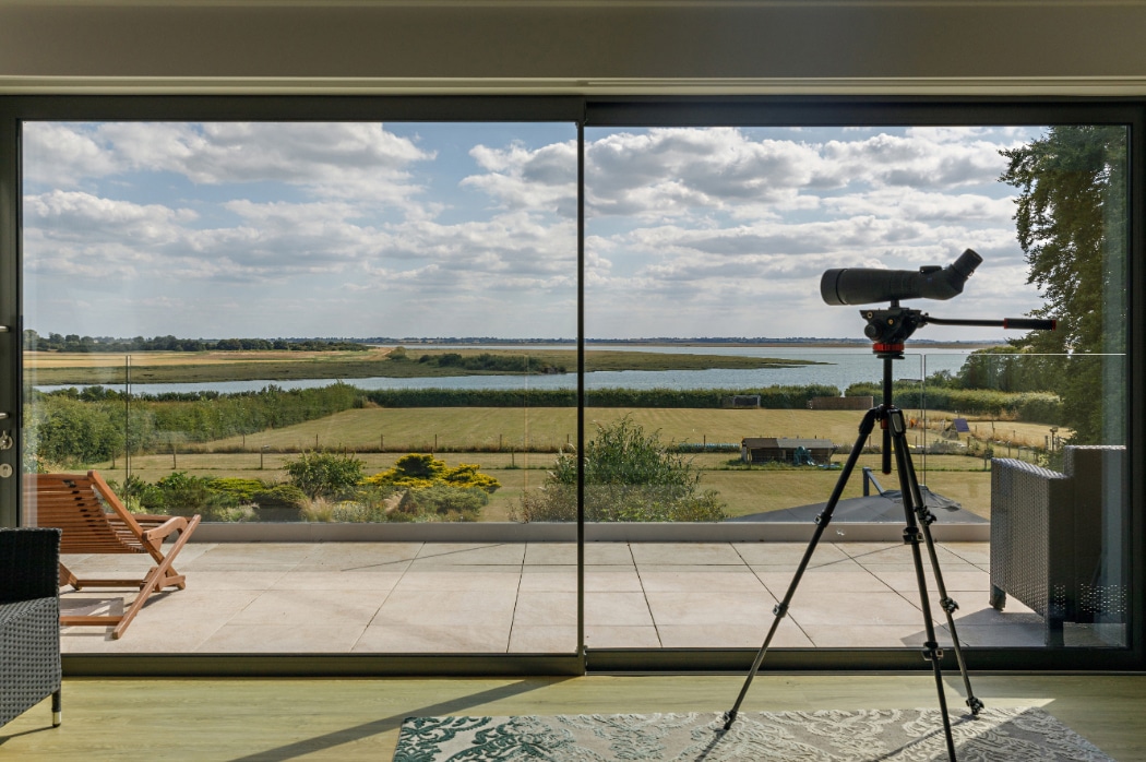 A telescope on a tripod is positioned indoors facing large glass doors, overlooking a sunny landscape with a river, fields, and a partly cloudy sky.