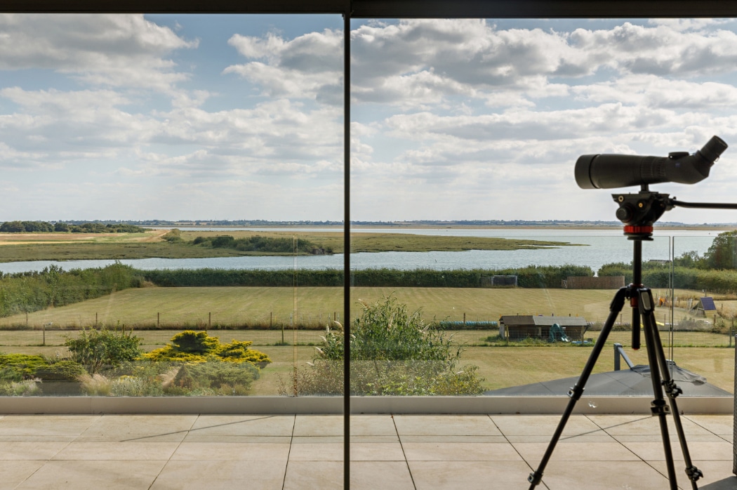 A large window overlooks a grassy garden, water, and distant fields under a partly cloudy sky; a spotting scope on a tripod stands indoors by the glass.