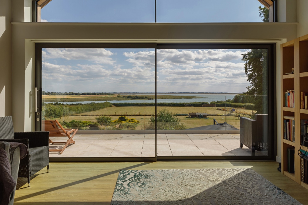 A large glass sliding door opens to a patio with loungers, overlooking a grassy field and a body of water under a partly cloudy sky.