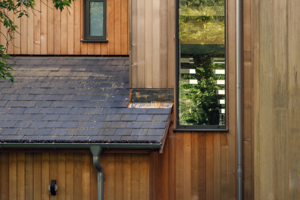 Close-up of a modern wooden house exterior, showing vertical timber panels, a slate roof, two windows, and guttering.