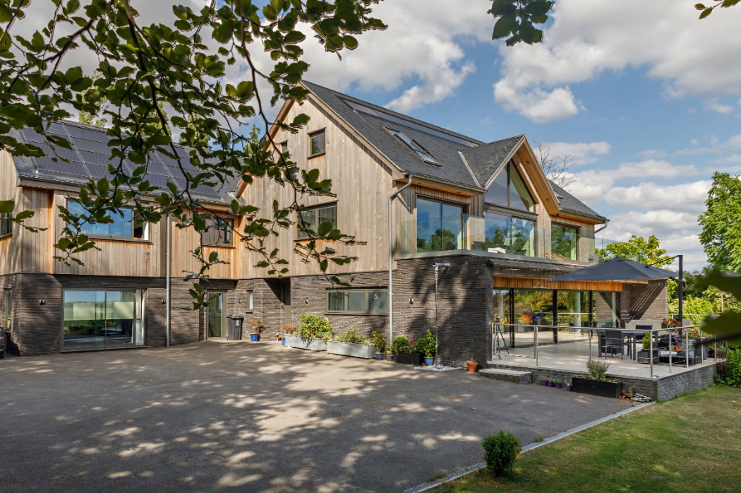 A modern two-storey house with wood and brick exterior, large windows, solar panels, and a spacious patio surrounded by trees under a partly cloudy sky.