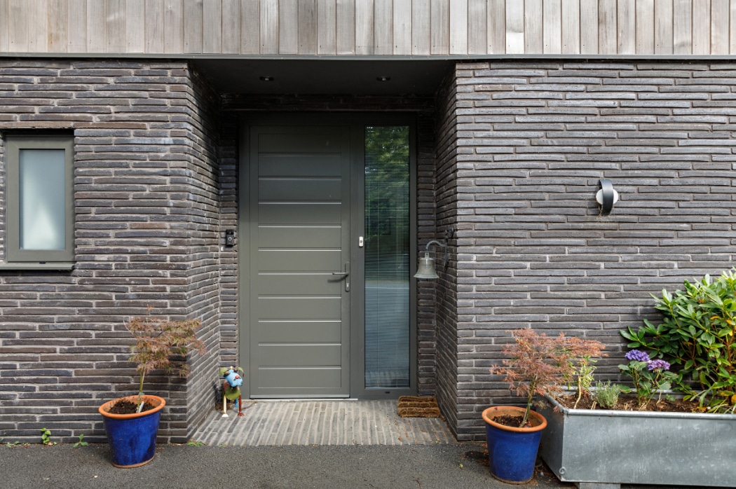 Modern grey front door set in a dark brick wall, flanked by two potted plants and a rectangular planter with greenery on the right.