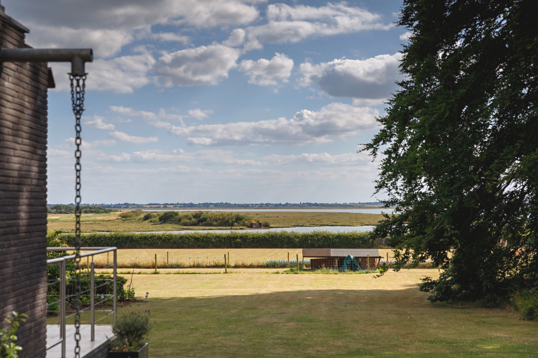 A grassy garden with a fence overlooks a river or lake, fields, and a cloudy sky; a tree and building are partially visible on the sides.