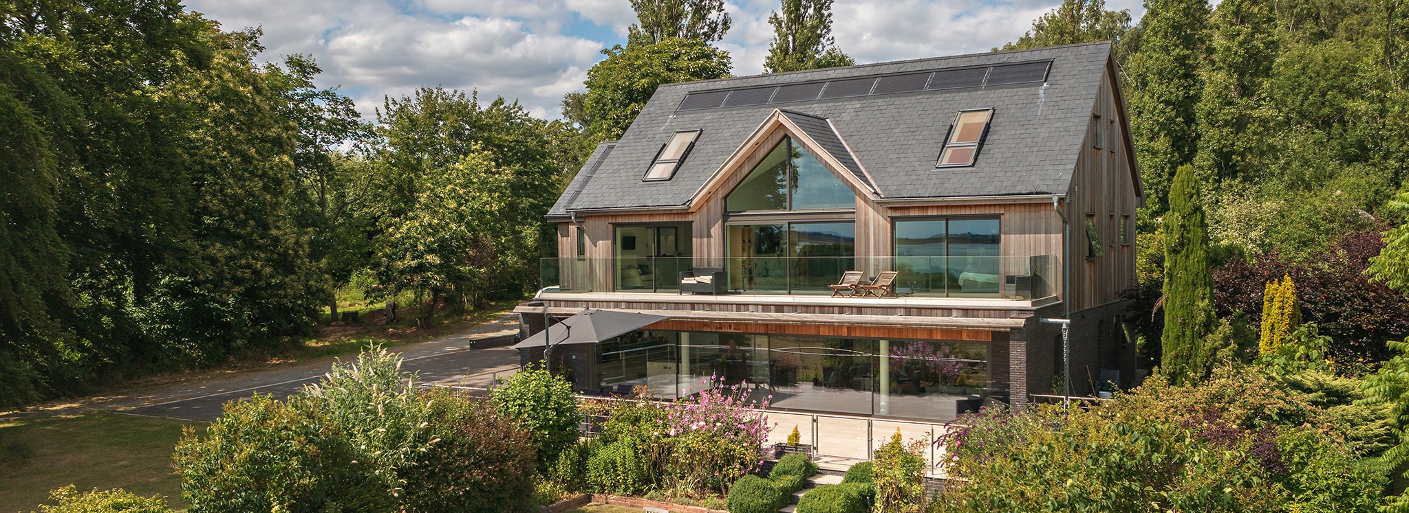 Modern two-storey house with large glass windows, solar panels on the roof, balcony seating, and surrounded by lush greenery and trees.