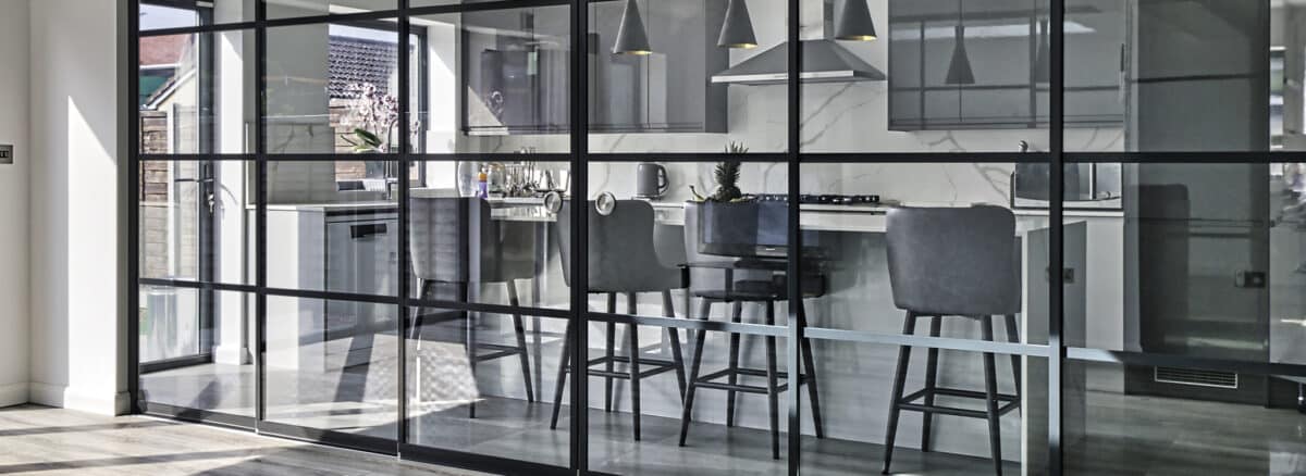 Modern kitchen with black-framed glass partition, high-top chairs at a counter, stainless steel appliances, and marble splashback.