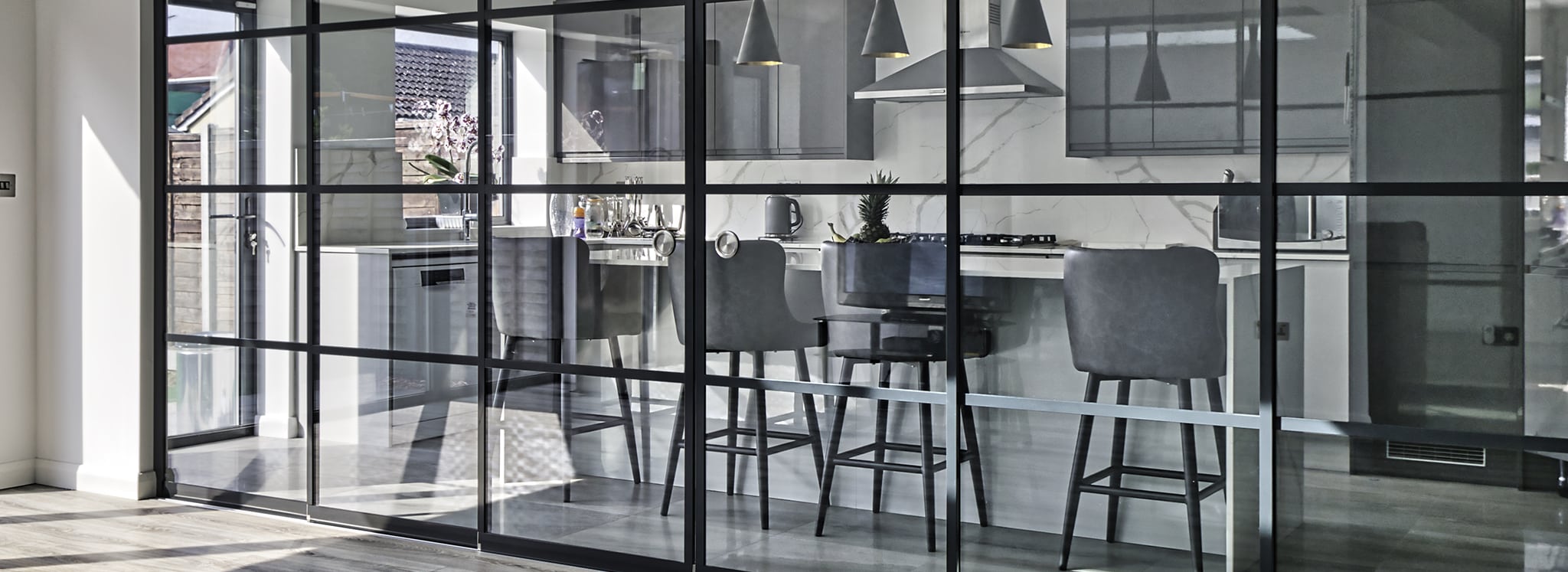 Modern kitchen with black-framed glass partition, high-top chairs at a counter, stainless steel appliances, and marble splashback.
