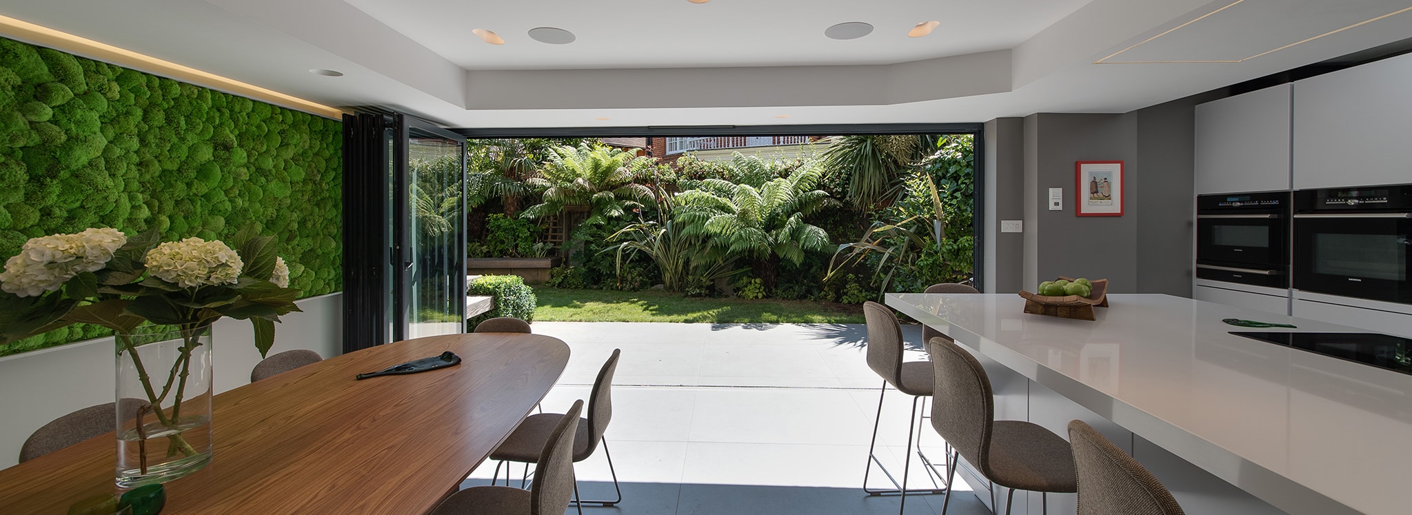 Modern kitchen and dining area with large glass doors open to a lush garden; features a living wall, integrated appliances, and a wooden dining table with chairs.