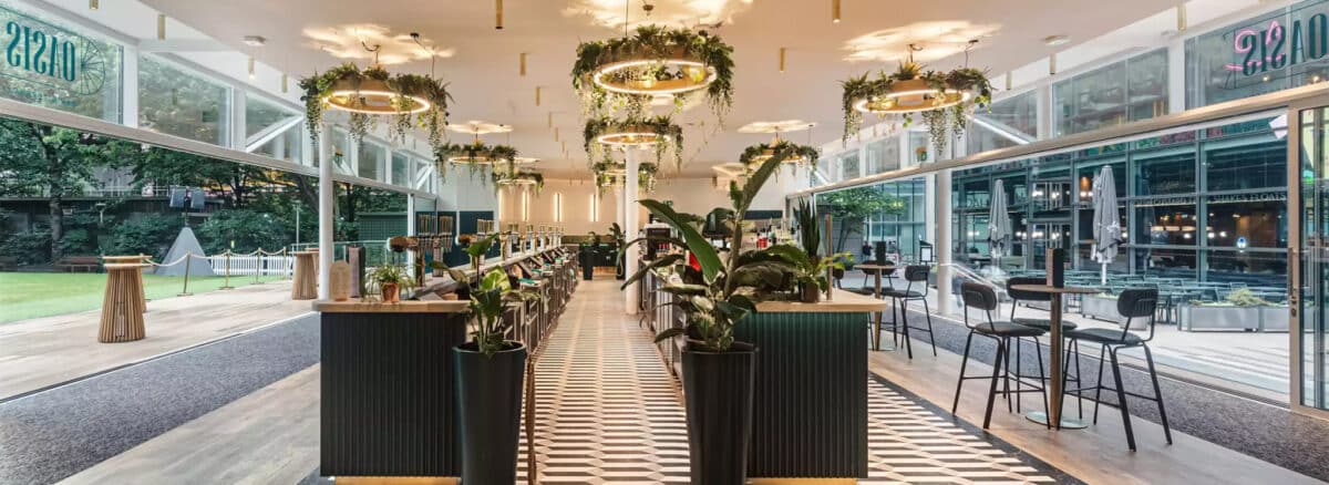 Modern restaurant interior with geometric floor tiles, bar seating, hanging ring lights with greenery, large windows, and decorative plants.