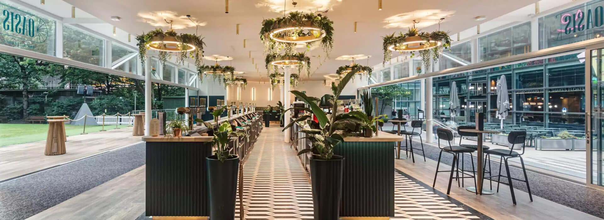 Modern restaurant interior with geometric floor tiles, bar seating, hanging ring lights with greenery, large windows, and decorative plants.