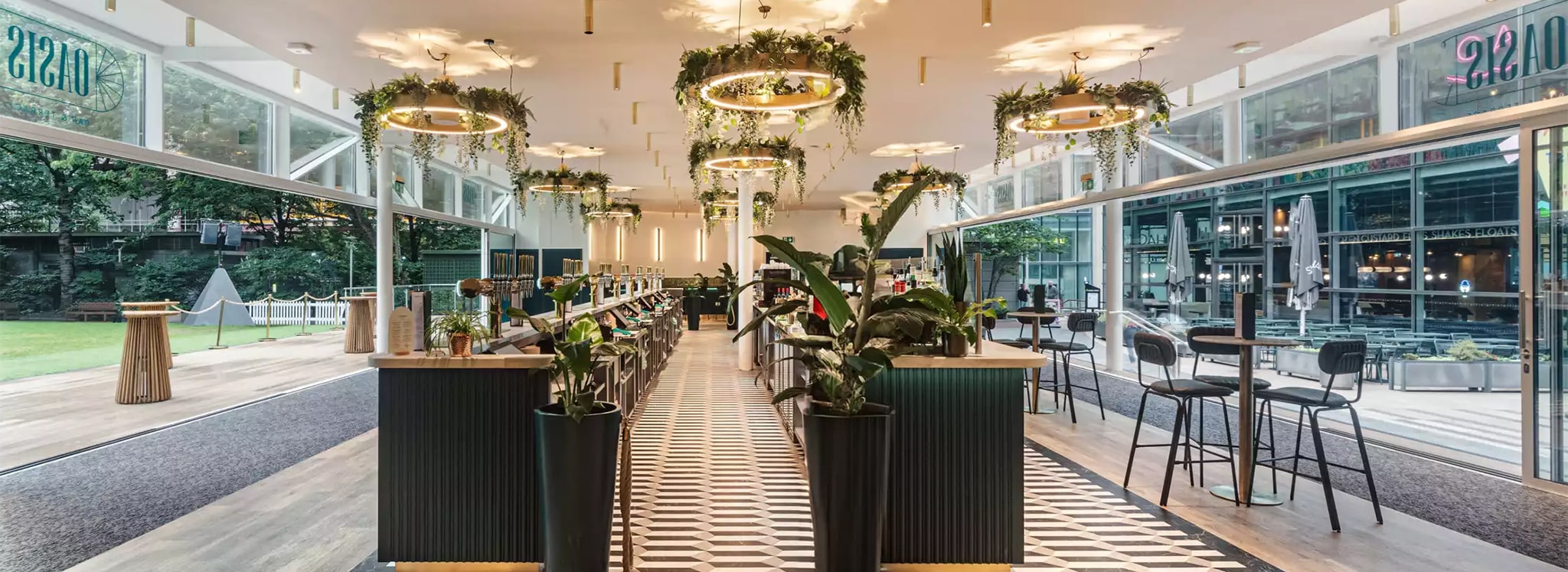 Modern restaurant interior with geometric floor tiles, bar seating, hanging ring lights with greenery, large windows, and decorative plants.