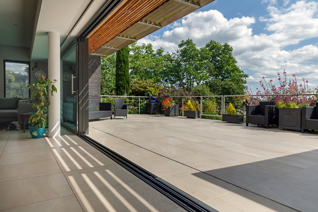 Spacious outdoor terrace with large tiles, potted plants, and metal railing, adjacent to an open modern living area under a partly cloudy sky.