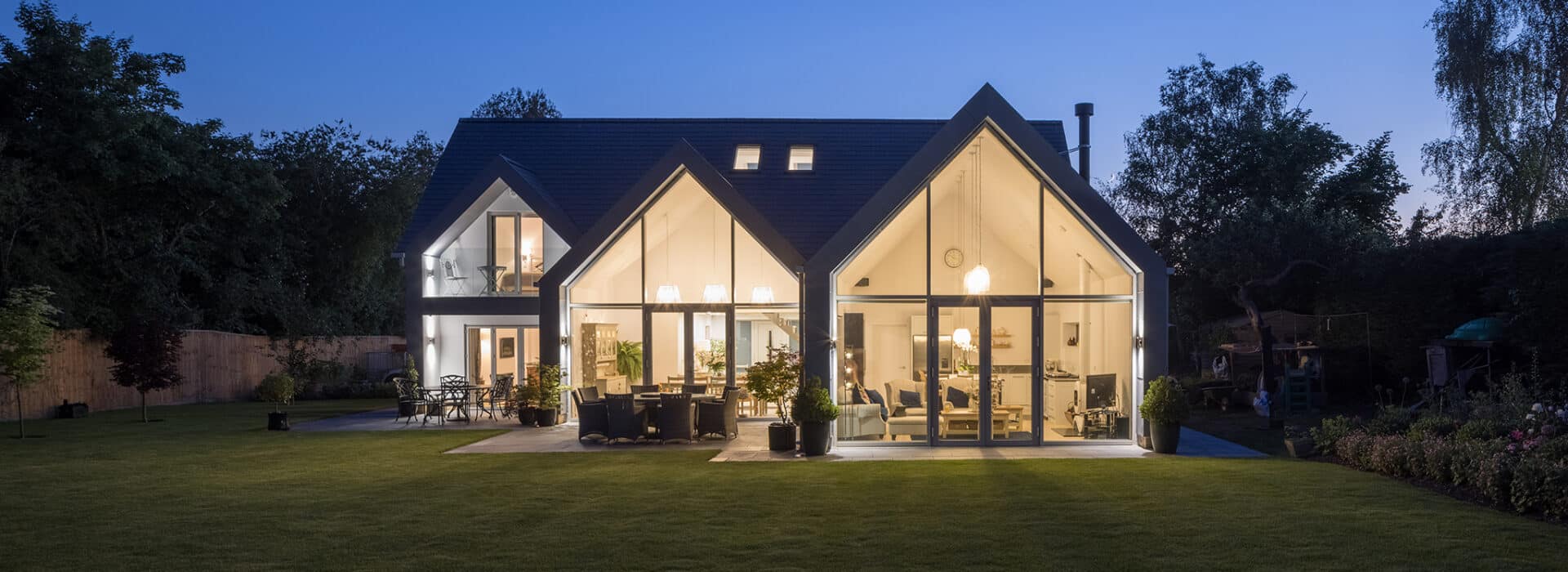Modern house with large glass windows and triangular roof, illuminated interior visible at twilight, surrounded by a lawn and trees.