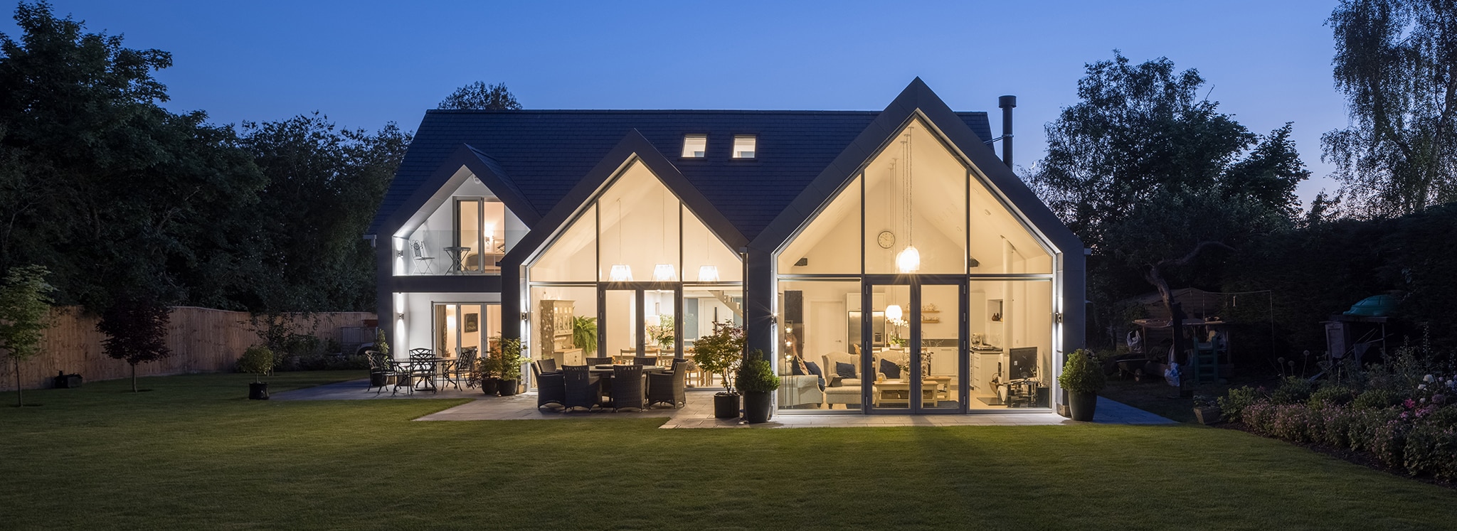 Modern house with large glass windows and triangular roof, illuminated interior visible at twilight, surrounded by a lawn and trees.