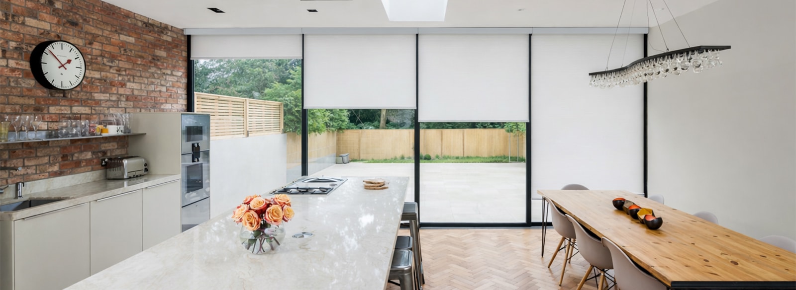 Modern kitchen and dining area with marble island, brick feature wall, large windows with blinds, wooden table, and outdoor patio view.
