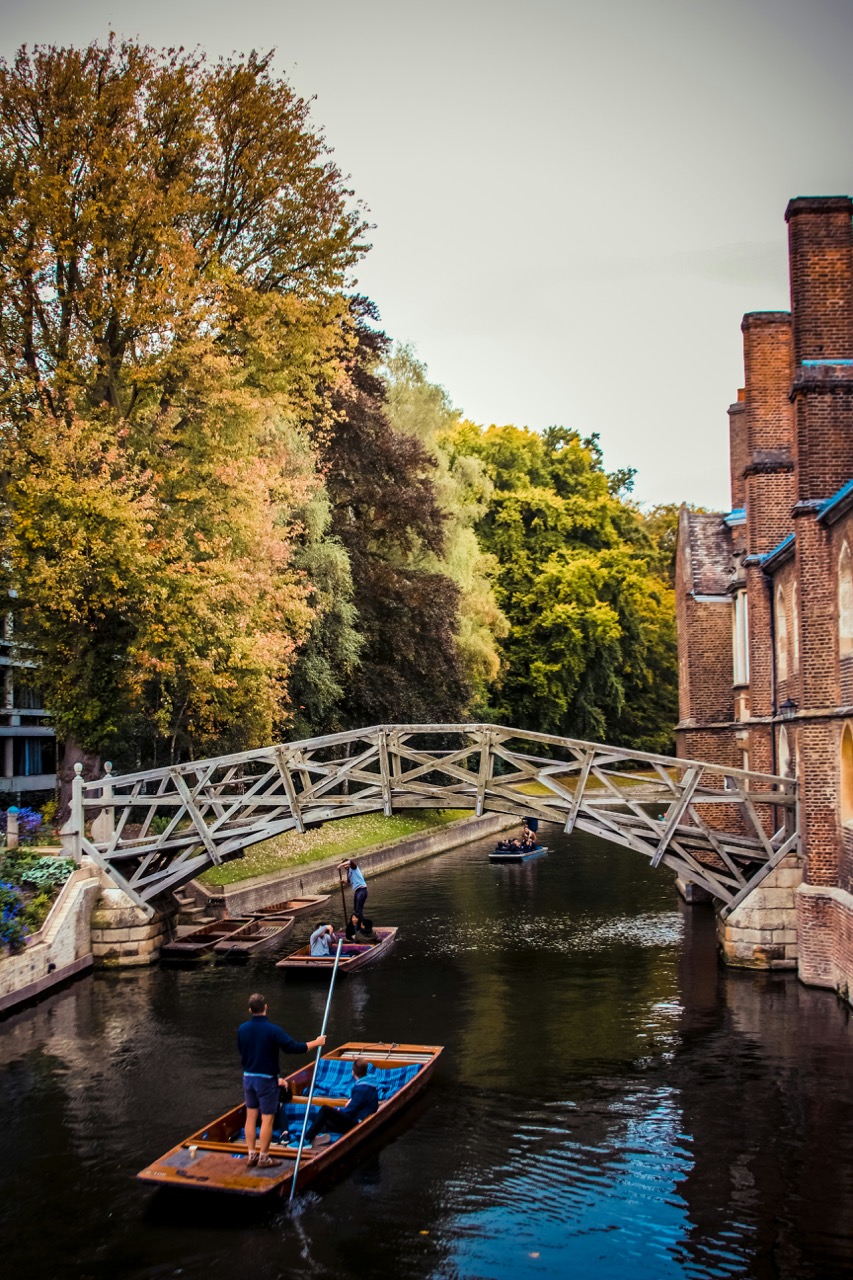 People are punting boats under a wooden bridge on a river, surrounded by trees and historic brick buildings.