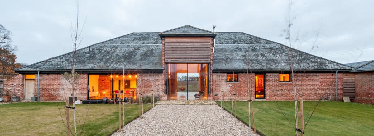 A modern barn conversion with large glass doors, brick walls, and a slate roof, with a gravel path leading to the entrance and neatly trimmed lawns on either side.