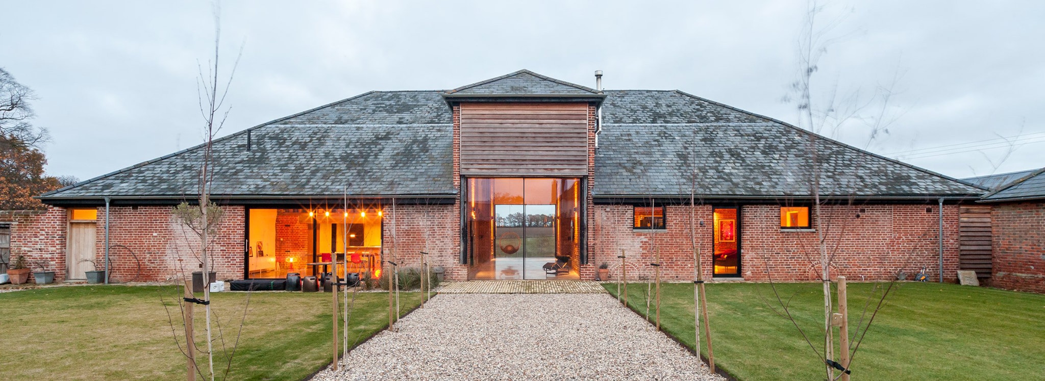 A modern barn conversion with large glass doors, brick walls, and a slate roof, with a gravel path leading to the entrance and neatly trimmed lawns on either side.