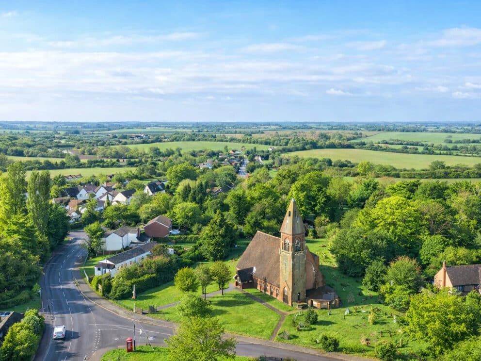 Aerial view of a rural village near the IDSystems Essex showroom, with a church, houses, trees, and green fields under a partly cloudy blue sky.
