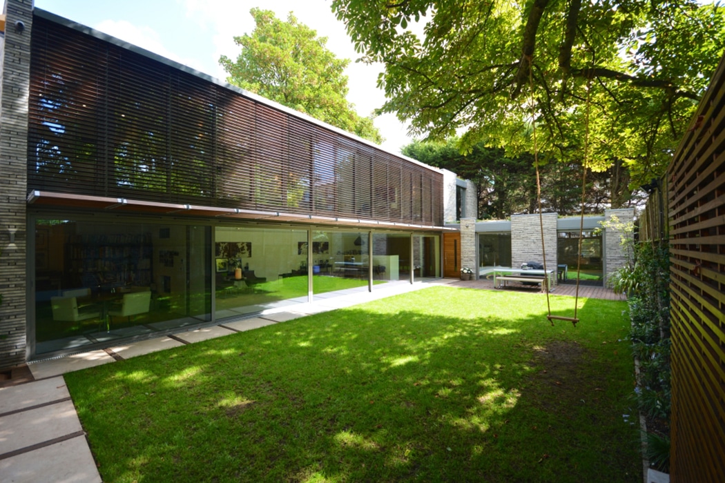 Modern two-storey house with large glass windows and sliding doors facing a green lawn and shaded by trees, featuring horizontal timber slats for privacy.