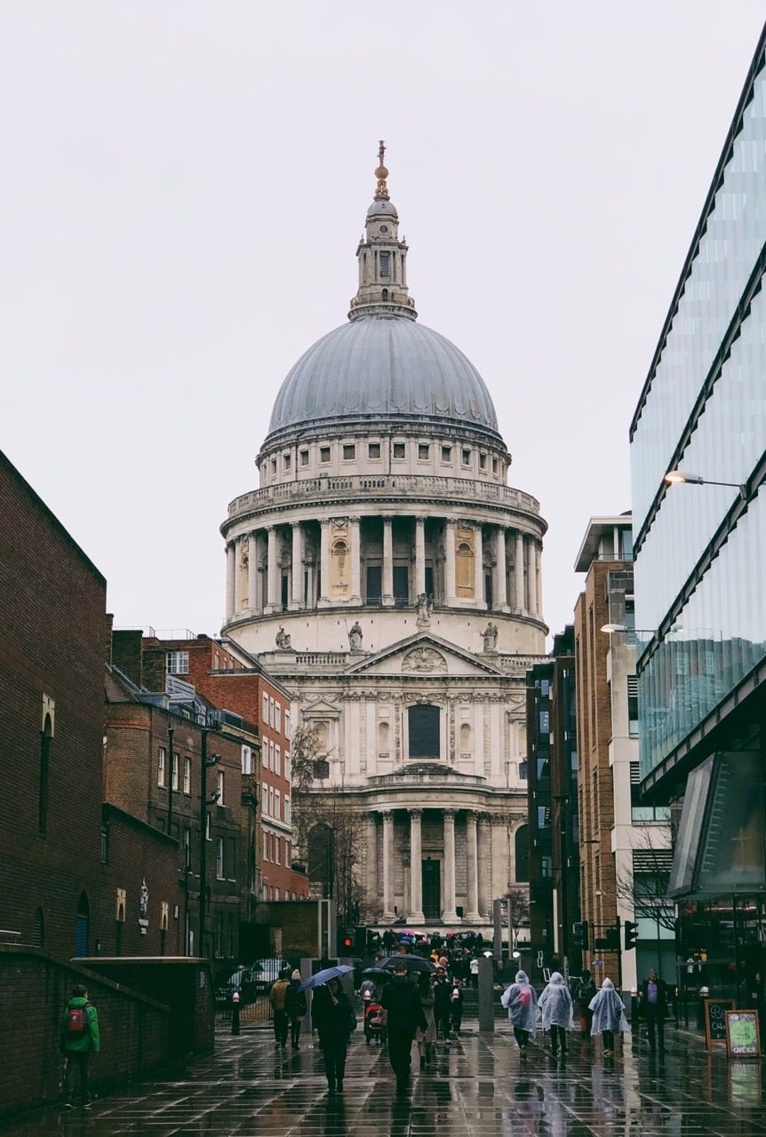 People walk on a rainy street towards St Paul’s Cathedral in London, with umbrellas visible and modern buildings flanking the historic structure.