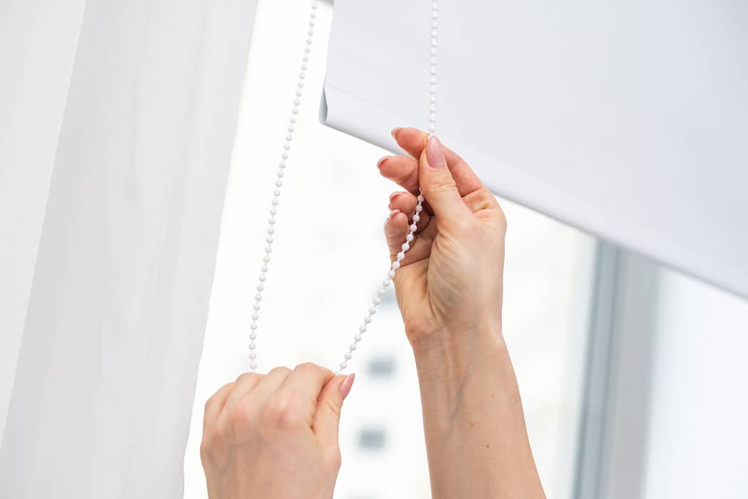 A person pulls a beaded chain to adjust a white roller blind near a window.